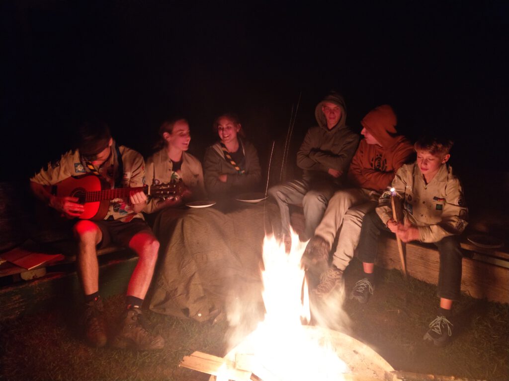Die Pfadis sitzen um das Lagerfeuer, eine Person spielt Gitarre und die Anderen singen. 