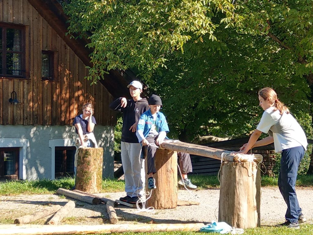Die Jugendlichen sind gerade dabei die Brücke zu bauen. Die Brücke besteht auf dem Bild aus drei Baumstümpfen als Stützen, wobei zwischen zweien gerade eine Lattung zum hinübergehen mit Seilen befestigt wird. 